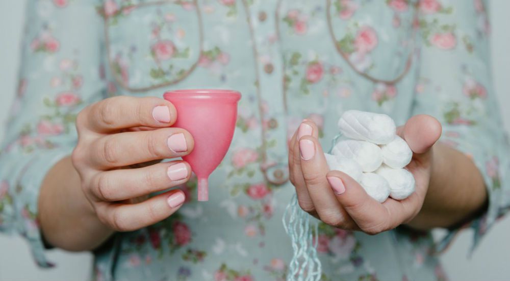 une femme tenant dans sa main droite une coupe menstruelle et dans la main gauche des tampons jetables