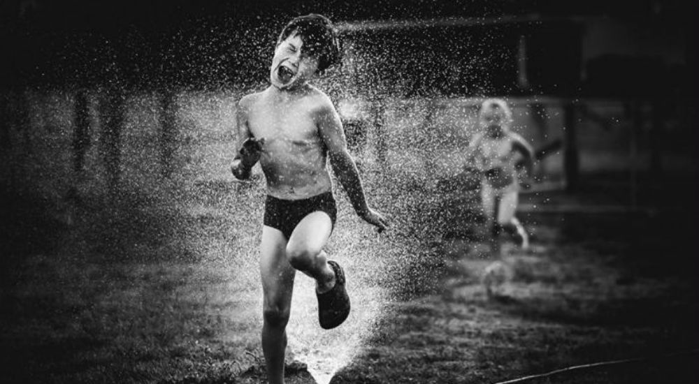 Un petit garçon avec un grand sourire court au dessus d'un jet d'eau pendant que ses petites soeurs le regardent