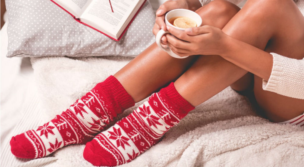 une femme boit une tisane chaude, lit un livre et porte des chaussettes de Noël